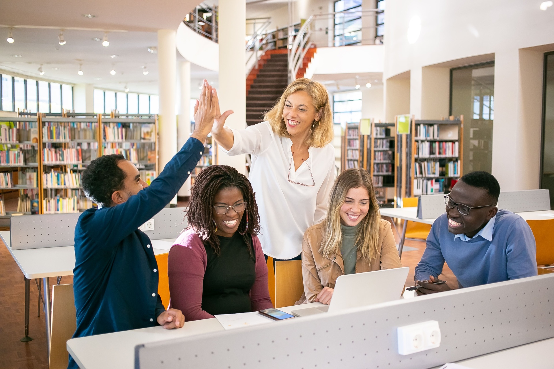 Students high-fiving a mentor while working on a laptop in a library